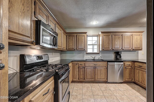 kitchen with brown cabinetry, stainless steel appliances, a textured ceiling, a sink, and light tile patterned flooring