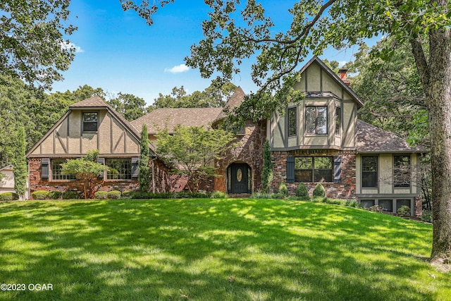 english style home featuring a shingled roof, brick siding, stucco siding, a front lawn, and a chimney