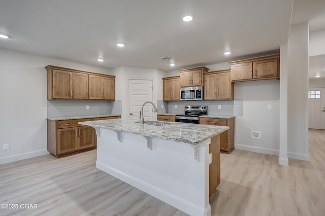 kitchen featuring appliances with stainless steel finishes, an island with sink, light hardwood / wood-style floors, light stone counters, and a breakfast bar