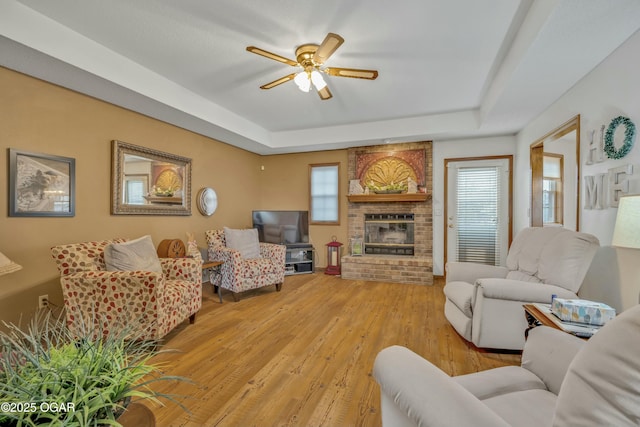 living room with ceiling fan, a raised ceiling, light wood-type flooring, and a brick fireplace