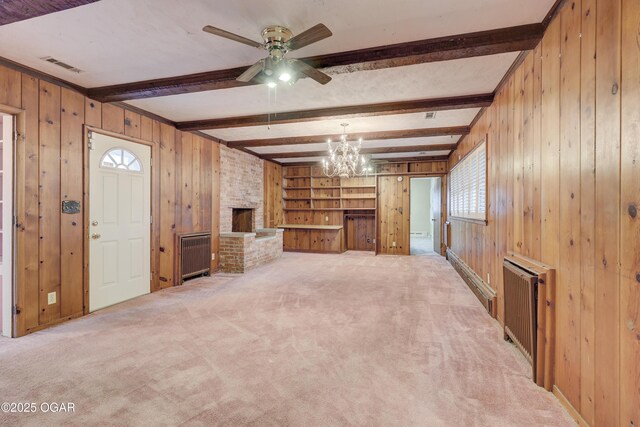 unfurnished living room featuring beamed ceiling, ceiling fan with notable chandelier, a brick fireplace, and wood walls