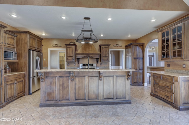 kitchen with an island with sink, appliances with stainless steel finishes, decorative light fixtures, light stone counters, and custom range hood