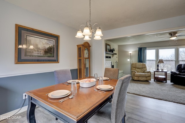 dining space featuring wood-type flooring, ceiling fan with notable chandelier, and a brick fireplace