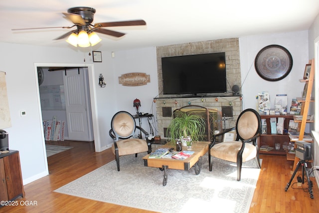 living room featuring baseboards, wood finished floors, ceiling fan, and a brick fireplace