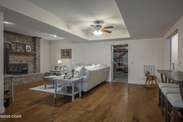 living room with ceiling fan, dark wood-type flooring, a fireplace, and a tray ceiling