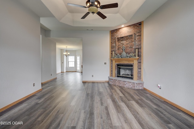 unfurnished living room featuring a brick fireplace, wood finished floors, a raised ceiling, baseboards, and ceiling fan with notable chandelier