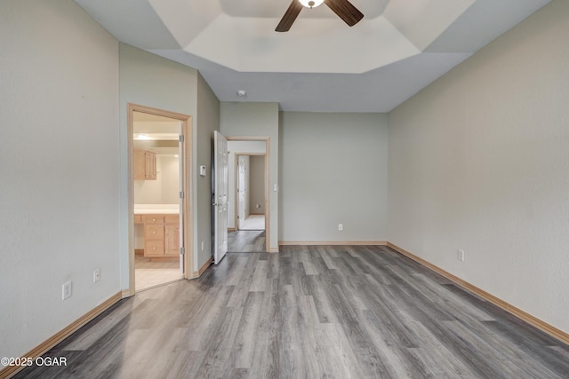 unfurnished bedroom with ceiling fan, wood finished floors, baseboards, ensuite bathroom, and a tray ceiling