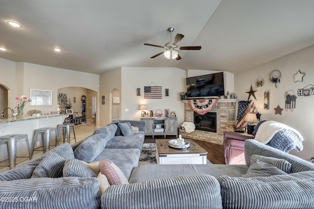 living area with arched walkways, lofted ceiling, ceiling fan, wood finished floors, and a brick fireplace