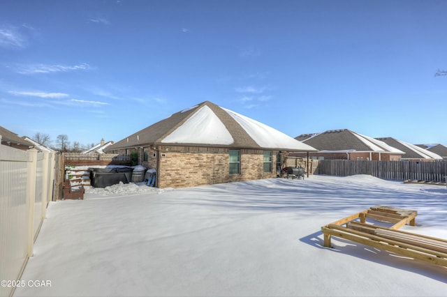 snow covered back of property featuring a hot tub, a fenced backyard, and brick siding