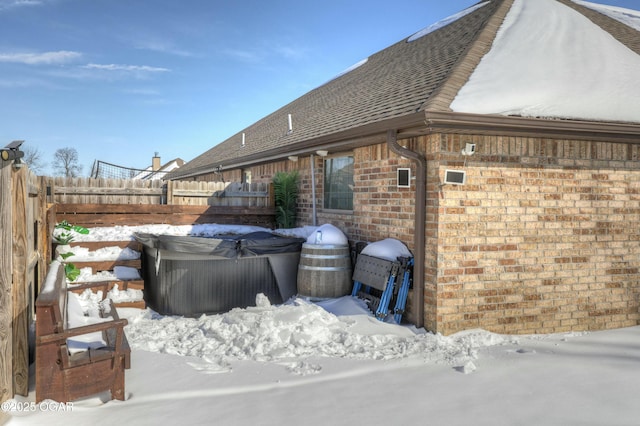 view of snowy exterior featuring a hot tub, fence, roof with shingles, and brick siding