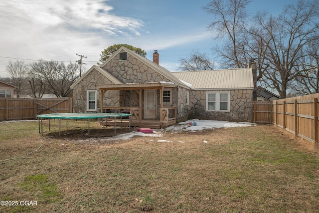 rear view of property featuring a trampoline, a fenced backyard, stone siding, and a chimney