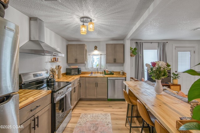 kitchen with appliances with stainless steel finishes, a sink, butcher block countertops, light wood-type flooring, and wall chimney exhaust hood