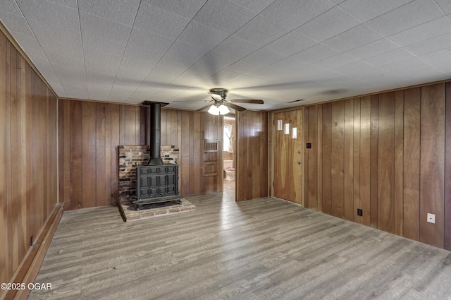 unfurnished living room featuring a ceiling fan, wood finished floors, a wood stove, and wooden walls