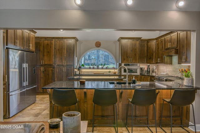 kitchen with a sink, appliances with stainless steel finishes, backsplash, dark stone countertops, and a kitchen bar