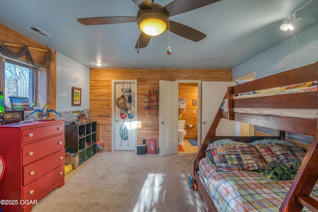 carpeted bedroom with a ceiling fan, wooden walls, visible vents, and connected bathroom