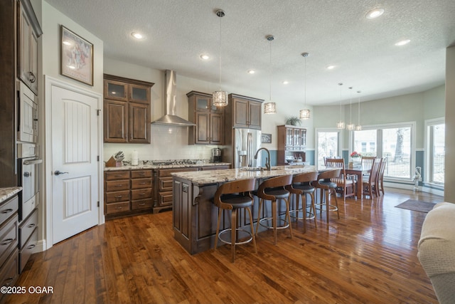 kitchen featuring tasteful backsplash, glass insert cabinets, wall chimney range hood, a kitchen bar, and stainless steel appliances