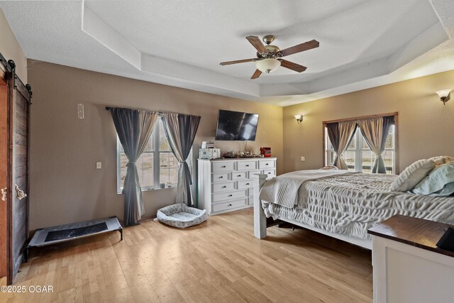 bedroom featuring a barn door, a raised ceiling, light wood-style flooring, and a ceiling fan