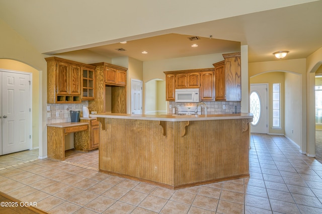 kitchen featuring light tile patterned floors, arched walkways, white microwave, a peninsula, and light countertops