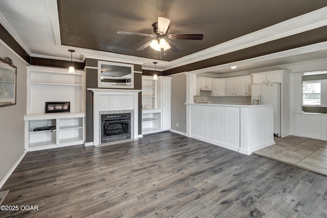 unfurnished living room featuring ceiling fan, dark wood-style flooring, a tray ceiling, a tiled fireplace, and crown molding