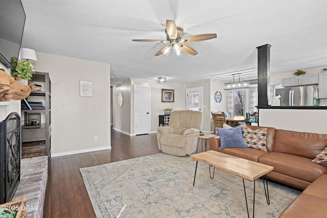 living area featuring dark wood-style floors, a brick fireplace, baseboards, and a ceiling fan