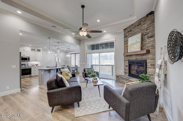 living area featuring a ceiling fan, baseboards, ornamental molding, a stone fireplace, and light wood-style floors