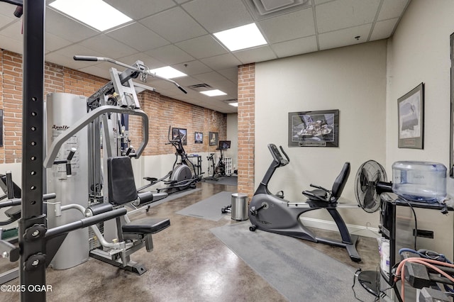 exercise room with a paneled ceiling, visible vents, brick wall, and baseboards