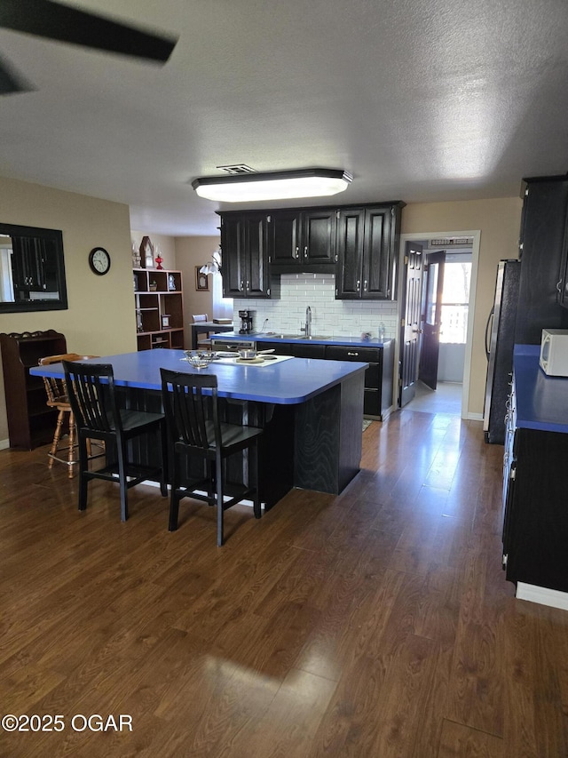 kitchen featuring dark cabinetry, dark wood finished floors, an island with sink, freestanding refrigerator, and tasteful backsplash