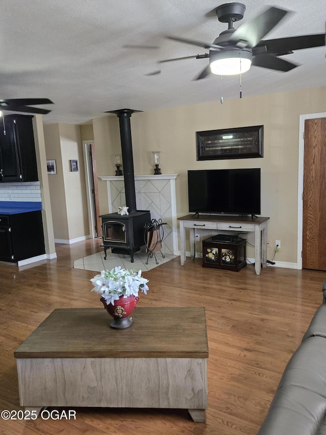 living area featuring wood finished floors, a ceiling fan, baseboards, a wood stove, and a textured ceiling