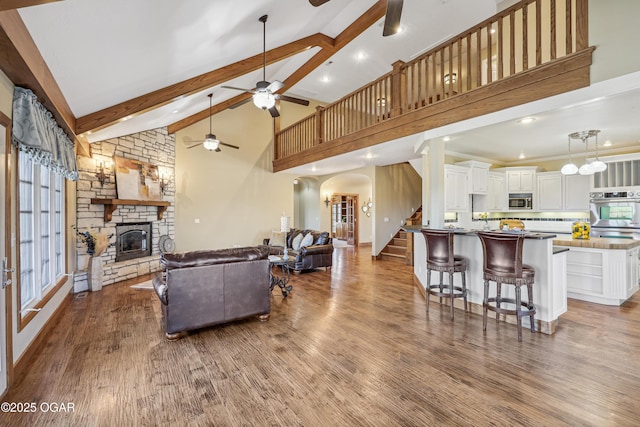 living room with a stone fireplace, beamed ceiling, a ceiling fan, and wood finished floors