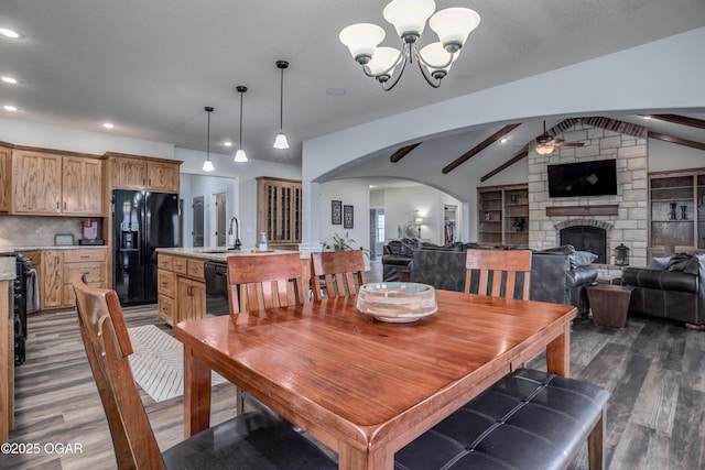 dining room with dark wood-style floors, lofted ceiling with beams, a fireplace, arched walkways, and ceiling fan with notable chandelier