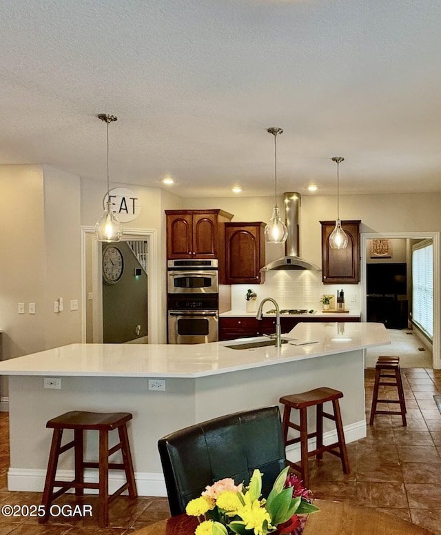 kitchen featuring wall chimney exhaust hood, a sink, stainless steel double oven, light countertops, and a breakfast bar area