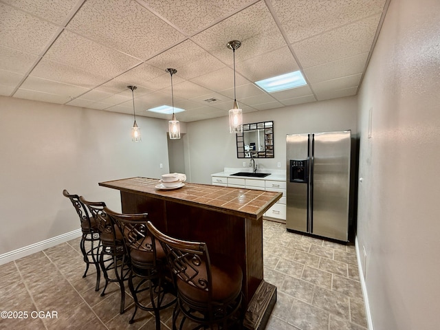 bar featuring stainless steel refrigerator with ice dispenser, a sink, a drop ceiling, hanging light fixtures, and baseboards