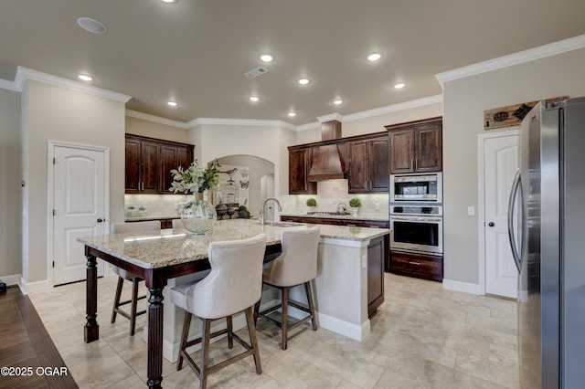 kitchen featuring appliances with stainless steel finishes, a kitchen island with sink, arched walkways, decorative backsplash, and a kitchen bar