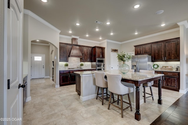 kitchen featuring custom exhaust hood, tasteful backsplash, a large island with sink, arched walkways, and appliances with stainless steel finishes