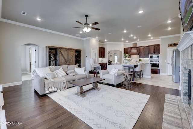 living room featuring wood-type flooring, ornamental molding, a brick fireplace, ceiling fan, and arched walkways
