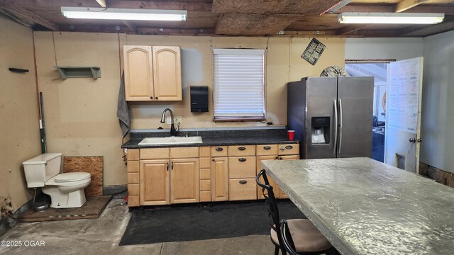 kitchen with a sink, concrete floors, light brown cabinets, dark countertops, and stainless steel refrigerator with ice dispenser