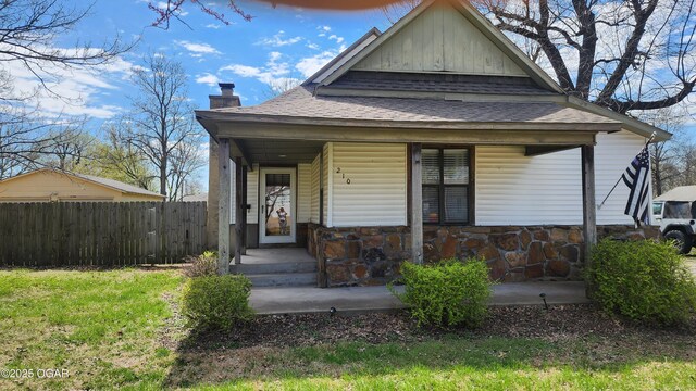 view of front facade with stone siding, fence, a chimney, covered porch, and a shingled roof