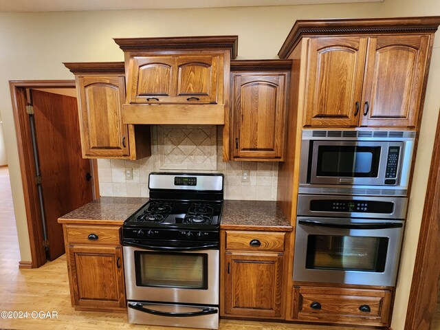 305 E Maple St, Liberal, MO 64762 - kitchen featuring brown cabinets, stainless steel appliances, light wood-type flooring, backsplash, and premium range hood