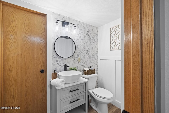 half bathroom featuring wood finished floors, toilet, wallpapered walls, and vanity