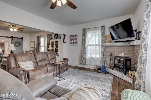 living area featuring a wood stove, wood finished floors, baseboards, a barn door, and a ceiling fan