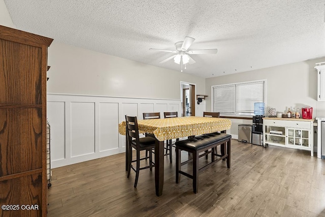 dining space with a decorative wall, a wainscoted wall, wood finished floors, a textured ceiling, and a ceiling fan