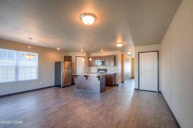 kitchen featuring stainless steel appliances, a kitchen bar, an island with sink, dark wood-style flooring, and baseboards