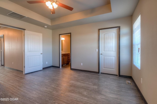 unfurnished bedroom with wood finished floors, baseboards, a raised ceiling, connected bathroom, and ceiling fan