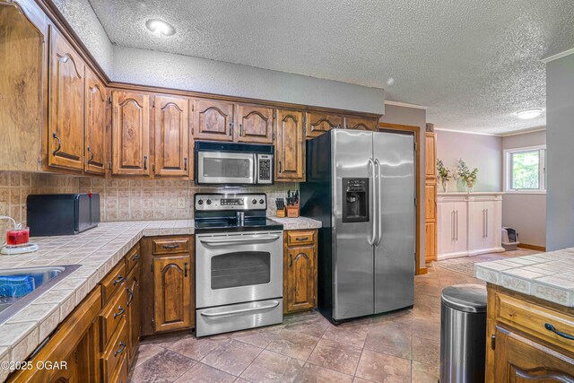 kitchen with appliances with stainless steel finishes, tile countertops, a textured ceiling, backsplash, and brown cabinetry