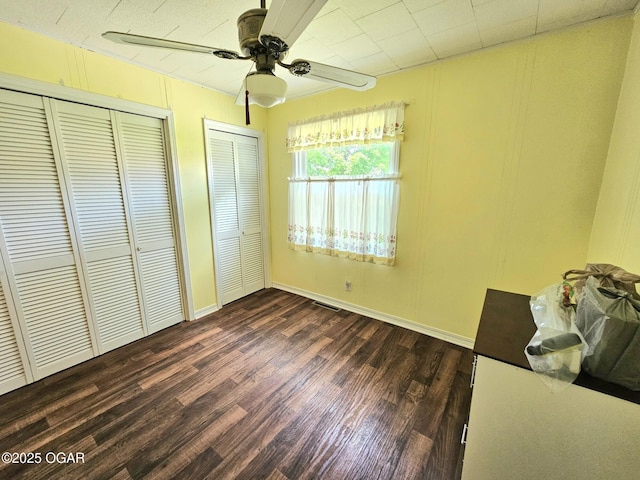 unfurnished bedroom featuring dark wood-style flooring, two closets, a ceiling fan, and baseboards