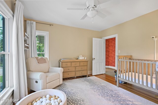 bedroom featuring wood finished floors, a nursery area, ceiling fan, and baseboards