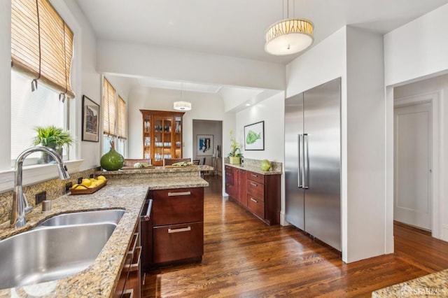 kitchen with a sink, dark wood finished floors, light stone countertops, hanging light fixtures, and stainless steel built in fridge