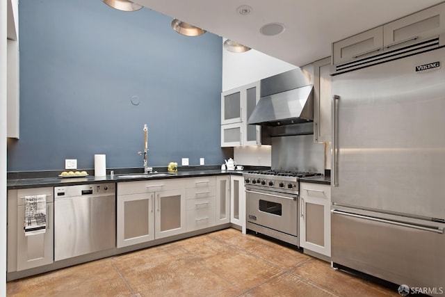 kitchen with high end appliances, dark countertops, glass insert cabinets, a sink, and wall chimney range hood