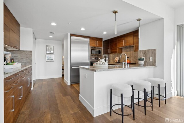 kitchen featuring dark wood-type flooring, built in appliances, a peninsula, a kitchen bar, and a sink