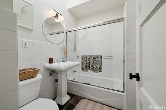 bathroom featuring enclosed tub / shower combo, toilet, wainscoting, and tile walls